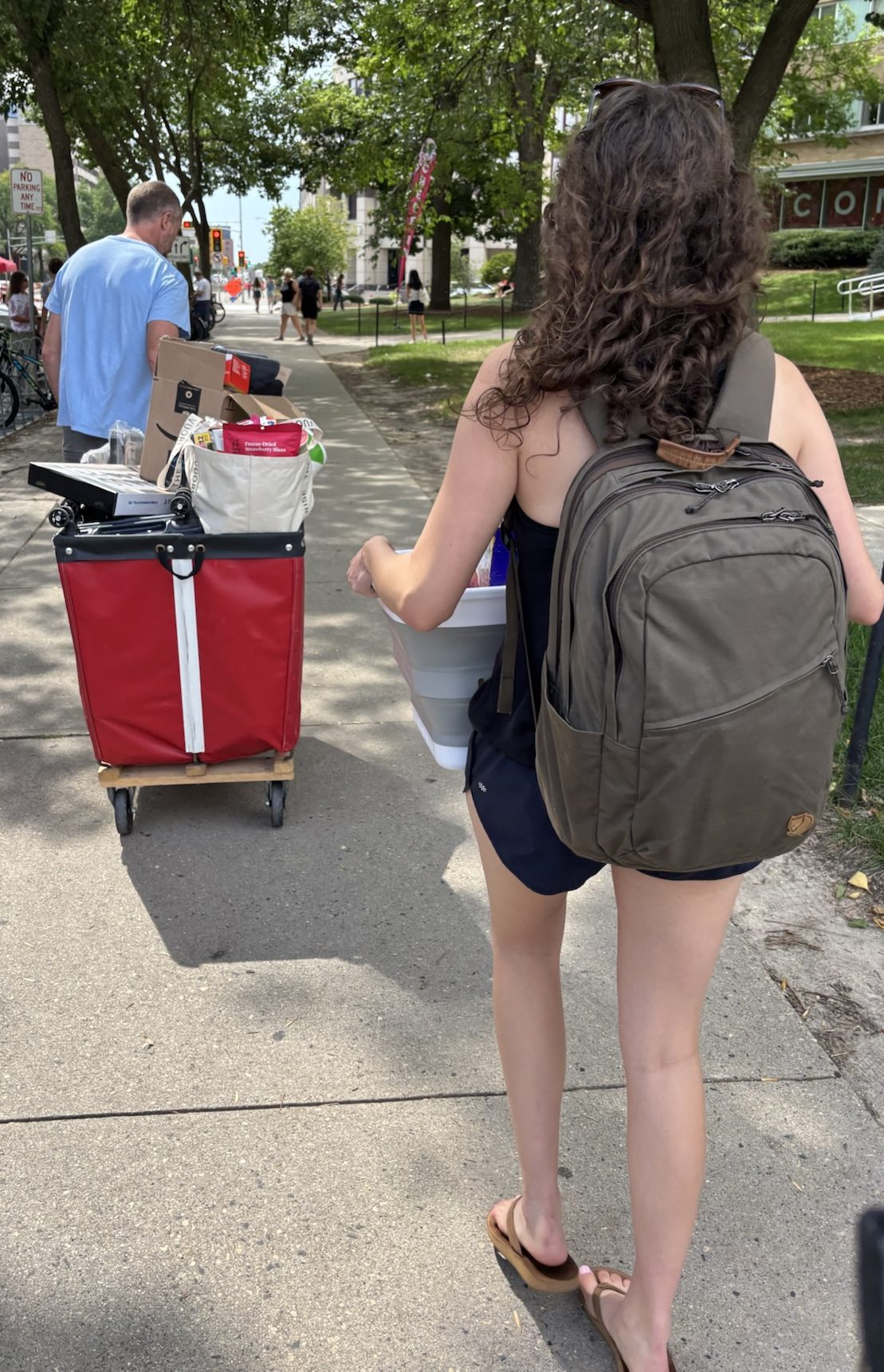 A dad wheeling a red cart of dorm stuff and a daughter wearing a backpack and carrying a basket of dorm stuff on a college campus